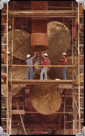Workers stand alongside an enormous ship propeller.