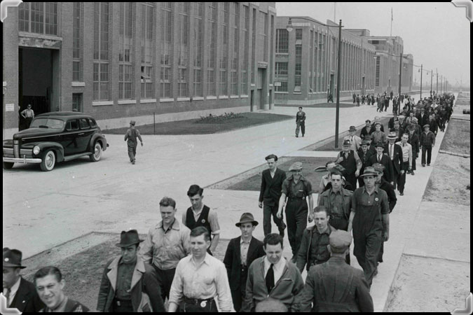 Photographie en noir et blanc montrant plusieurs travailleurs sortant de l'usine et une auto ancienne en arrière-plan.