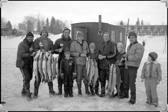 Several adults and children in front of their ice fishing cabin, showing off the numerous pike they have landed.
